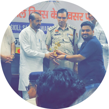 Man in a dark t-shirt receiving a trophy on stage during Skill Saathi Award ceremony from a dignitary in white kurta alongside a police officer.