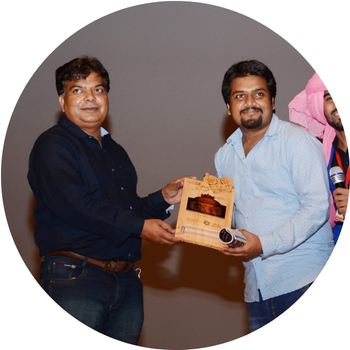Man in a light blue shirt receiving a decorative wooden memento plaque on stage from a guest in a dark shirt.